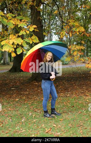 Attraktive Frauen aus Kaukasien mittleren Alters in Belleisle Park, Ayr, Ayrshire, Schottland, Großbritannien Genießen Sie die Herbstfarben in Jeans und schwarzem Wolltop und tragen einen bunten Regenbogen-Regenschirm mit Brolly Stockfoto