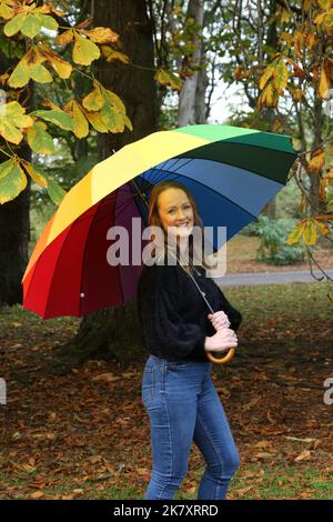 Attraktive Frauen aus Kaukasien mittleren Alters in Belleisle Park, Ayr, Ayrshire, Schottland, Großbritannien Genießen Sie die Herbstfarben in Jeans und schwarzem Wolltop und tragen einen bunten Regenbogen-Regenschirm mit Brolly Stockfoto