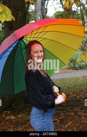 Attraktive Frauen aus Kaukasien mittleren Alters in Belleisle Park, Ayr, Ayrshire, Schottland, Großbritannien Genießen Sie die Herbstfarben in Jeans und schwarzem Wolltop und tragen einen bunten Regenbogen-Regenschirm mit Brolly Stockfoto