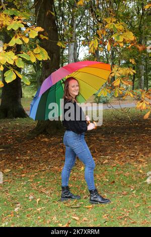 Attraktive Frauen aus Kaukasien mittleren Alters in Belleisle Park, Ayr, Ayrshire, Schottland, Großbritannien Genießen Sie die Herbstfarben in Jeans und schwarzem Wolltop und tragen einen bunten Regenbogen-Regenschirm mit Brolly Stockfoto