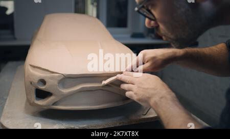 Die Hände eines männlichen Designers arbeiten an der Skulptur eines Autos mit Werkzeugen zur Bildhauerei. Glättet die Oberfläche des unfertigen Modells in einer Werkstatt. Automobilindustrie. Stockfoto