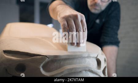 Erfahrene Autodesigner arbeiten an der Skulptur eines Prototypen-Automodells aus rohem Lehm mit Bildhauerwerkzeugen. Autodesigner mit gerahmten Gläsern arbeiten eng zusammen, um die Oberfläche der Motorhaube zu glätten. Stockfoto