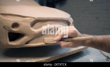 Die Hände eines männlichen Designers arbeiten an der Skulptur eines Autos mit Werkzeugen zur Bildhauerei. Glättet die Oberfläche des unfertigen Modells in einer Werkstatt. Automobilindustrie. Stockfoto