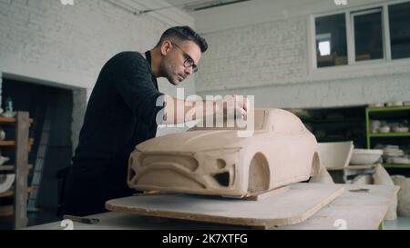 Ein Keramiker arbeitet eng an den Designdetails einer unfertigen umweltfreundlichen Prototypen-Skulptur eines Elektroautos aus rohem Lehm. Eine Designerin mit Brille und Schürze arbeitet in einer Keramikwerkstatt. Stockfoto