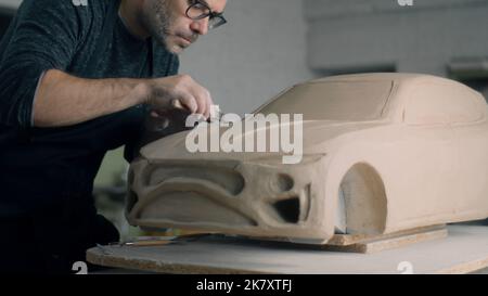 Der erfahrene Autodesigner arbeitet am Prototypenmodell eines Autos mit Holzformwerkzeugen, um die Motorhaubenoberfläche zu glätten. Unvollendetes Automodell auf einem Holztisch platziert. Stockfoto