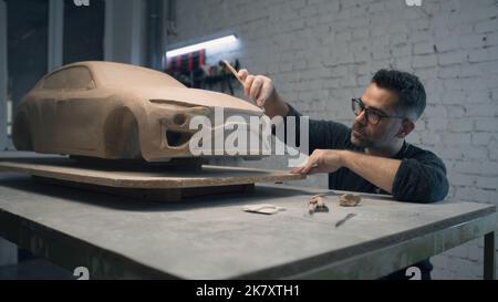 Senior Car Designer arbeitet an der Skulptur des umweltfreundlichen Auto-Prototyps mit Holzformwerkzeug, um die Oberfläche zu glätten und Details im Design zu schaffen. Handgemachte Auto Skulptur aus Ton. Stockfoto