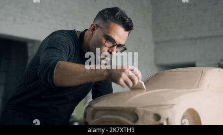 Der erfahrene Automobildesigner mit Schürze arbeitet an der Lehmskulptur des umweltfreundlichen Autos, indem er die Designdetails mithilfe eines Holzmodellierwerkzeugs schnitzt. Handgemachte Auto Skulptur aus Ton Stockfoto