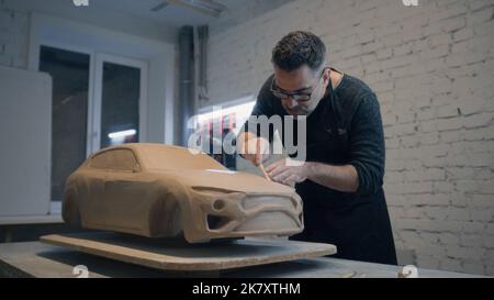 Erfahrene Designer arbeiten an der Skulptur des Autos aus rohem Lehm mit Holzformwerkzeug, um die Oberfläche zu glätten. Handgemachte Auto Skulptur aus Ton. Automobilindustrie. Stockfoto