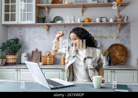 Hispanische Geschäftsfrau, die von zu Hause aus arbeitet und den Sieg und Erfolg feiert, hält die Hände hoch und schaut auf den Laptop, der gute Nachrichten liest. Stockfoto