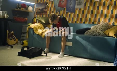 Kleiner Junge praciert Fußball in seinem Schlafzimmer. Junger begeisterter Fußballspieler vor dem Spiel. Ein moderner Raum mit Gitarre, Plakaten an der Wand und einem Schreibtisch mit Lampe. Stockfoto