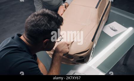 High-Angle-Aufnahme von zwei männlichen Automobildesignern, die an einem umweltfreundlichen Knetmasse-Modell des Autos mit Spezialwerkzeugen arbeiten. Zukünftiges Design von Elektroautos der neuen Generation. Arbeiten Sie in einem modernen Autodesignstudio Stockfoto