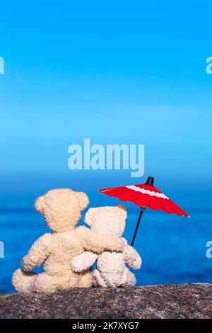 Teddybär-Freunde sitzen auf Stein am Meer Arm in Arm mit altem Sonnenschirm (Kopierplatz) Stockfoto