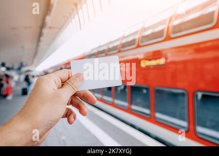 Kredit- oder Prepaid-Transportkarte in der Hand des Passagiers vor dem Hintergrund eines modernen Doppelstockzugs mit hoher Geschwindigkeit auf der Bahnplattform Stockfoto