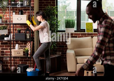 Hausfrau bewegen schwere Regale zu Hause afroamerikanischen Ehemann hilft Staubsaugen Boden. Multirassisches Paar, das im Frühjahr putzt und Möbel umzieht, um der Wohnung einen neuen Stil zu verleihen. Stockfoto
