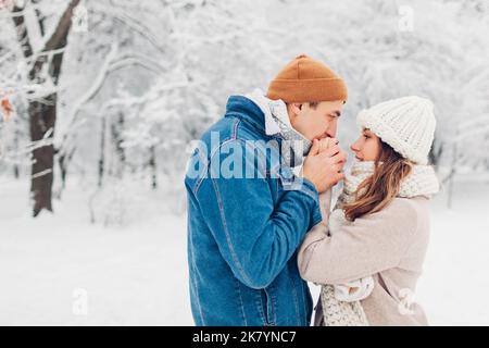 Mann wärmte die Hände der Frau mit seinem Atem im verschneiten kalten Winterpark. Liebevolles Paar, das im Freien läuft Stockfoto