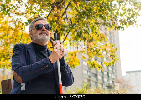 Blinder bärtiger grauhaariger reifer Mann mit dunkler Brille und einer Jacke, die einen Spazierstock hält und auf einer Bank im Park sitzt. Hochwertige Fotos Stockfoto