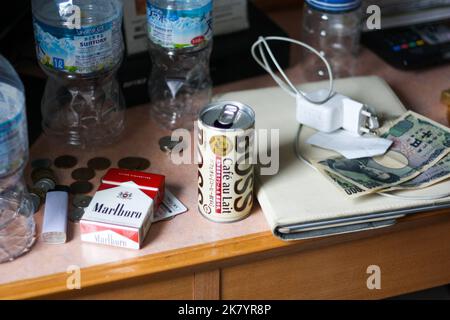 Unordentlicher kleiner Holzschreibtisch im Hotelzimmer mit Wasserplastikflaschen und japanischen Banknoten und Münzen und Zigarettenpackung, Kaffee in Dosen und Ladegerät. Stockfoto