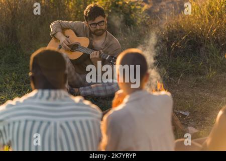 Kaukasischer Bärte kaufen in einer Brille, die vor seinen Freunden auf dem Gras sitzt und akustische Gitarre spielt. Außenaufnahme. Verschwommene Personen im Vordergrund. Hochwertige Fotos Stockfoto