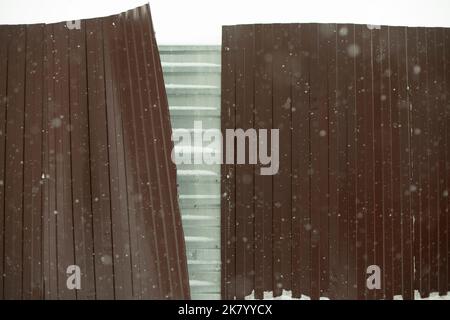 Zaun gebrochen. Stahlprofil im Wind. Der Zaun wird im Winter von Windböen weggerissen. Konstruktionsdetails. Stockfoto