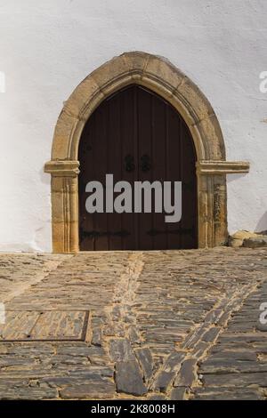 Weiß getünchtes Gebäude mit Eingangstür im gotischen Stil in einer Gasse, die mit Kopfsteinpflaster innerhalb der Mauern des Alten Schlosses in Monsaraz, Portugal, gepflastert ist. Stockfoto