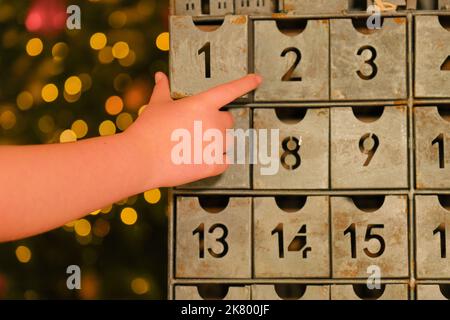 Metallischer Adventskalender im Vintage-Stil und Kinderhände in der Nähe des Weihnachtsbaums mit leuchtender Girlande auf verschwommenem Raumhintergrund. Weihnachtstraditionen und Stockfoto