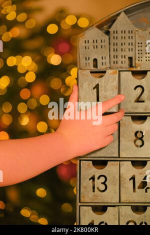 Metallischer Adventskalender und Kinderhände in der Nähe des Weihnachtsbaums mit glühender Girlande auf verschwommenem Raumhintergrund. Weihnachtstraditionen und -Symbole. Geschenke Stockfoto