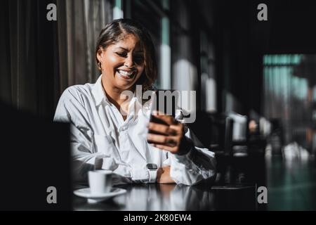 Ein Porträt einer lachenden, schönen und großen hispanischen Unternehmerin, die lacht, während sie einen Videoanruf mit einem Kollegen über das Smartphone führt, während sie sitzt Stockfoto