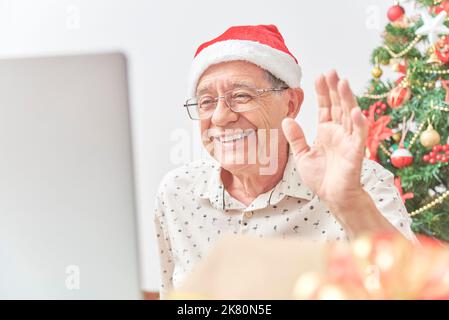 Ein älterer hispanischer Mann begrüßt seine Familie während der Weihnachtszeit per Videoanruf mit einem Laptop. Konzepte: Die Freude am Teilen während der Feiertage, die Verwendung von t Stockfoto