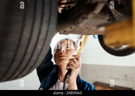 Mein Wissen über Autos ermöglicht es mir, Probleme viel schneller zu diagnostiziere. Eine Mechanikerin, die unter einem hochgelaufenen Auto arbeitet. Stockfoto