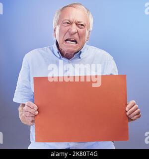 Mürrisch wird Sie mürrisch zeigen. Studioaufnahme eines älteren Mannes, der ein leeres Schild in der Hand hält und vor blauem Hintergrund unglücklich aussieht. Stockfoto