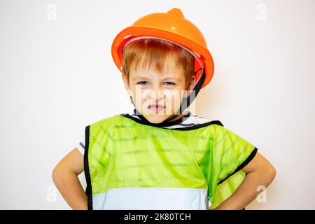 Der Junge im Helm und in der Weste ist empört. Ein kleiner Arbeiter. Der Junge ist ein Baumeister. Stockfoto