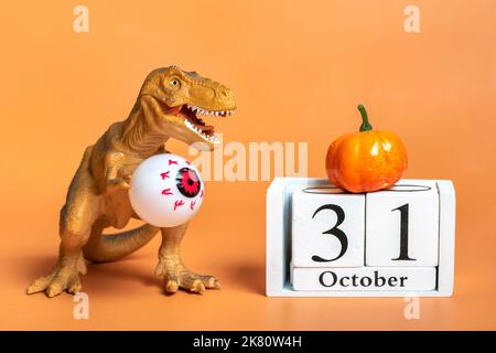 Spielzeugdinosaurier Tyrannosaurus mit Augen in den Pfoten, Kürbis, Kalenderdatum 31 Oktober isoliert auf orangefarbenem Hintergrund Grußkarte Happy Hallow Stockfoto