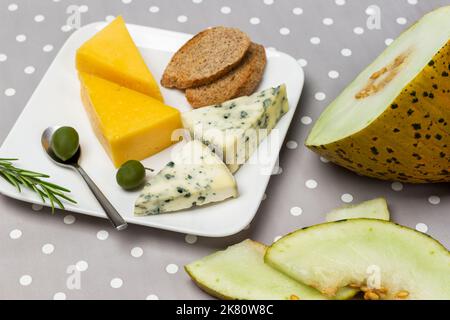 Melone, verschiedene Käsesorten und Oliven auf dem Teller. Draufsicht. Hintergrund mit grauem Punktmuster. Stockfoto