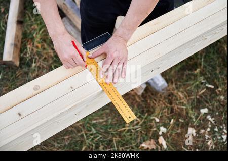 Mann Arbeiter Gebäude Holzrahmenhaus auf Pfahlfundament. Nahaufnahme des Zimmermanns mit Zentimeter zum Messen von Holzbohlen und zum Markieren mit Bleistift. Schreinerei-Konzept. Stockfoto