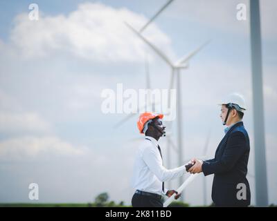 Afrikanischer Ingenieur-Mann im weißen Hemd schüttelt sich die Hände mit asiatischem Ingenieur im schwarzen Anzug mit Windturbinen im Hintergrund. Zwei Kollegen, die sich darüber austauschen Stockfoto