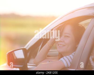 Schöne lächelnde asiatische Frau aus dem Fenster Blick weißes Auto bei Sonnenuntergang auf dem Feld in der Natur.Reise-und Urlaubskonzept Stockfoto