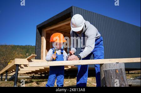 Vater mit Kleinkind Sohn Gebäude Holzrahmen Haus. Männliche Bauherren, die auf der Baustelle Nagel in die Planke schlagen, tragen an sonnigen Tagen Helm und blaue Overalls. Zimmerei- und Familienkonzept. Stockfoto