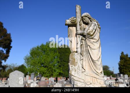 Alte Skulptur einer Frau, die eine Robe trägt, drapiert auf einem Friedhof über ein christliches Kreuz, mit Reihen von Gräbern und Bäumen im Hintergrund Stockfoto