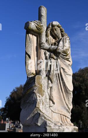 Abgenutzte und moosige Steinskulptur einer Frau, die eine Robe trug, während sie sich auf einem Kruzifix auf einem Friedhof stützte, mit hellblauem Himmel im Hintergrund Stockfoto