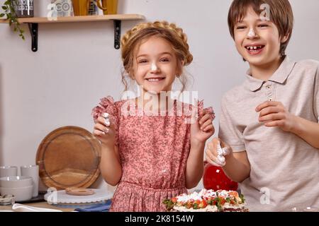 Kleine Freunde machen gemeinsam einen Kuchen in einer Küche gegen eine weiße Wand mit Regalen darauf. Stockfoto