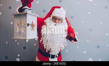 Weihnachtsmann, der Laterne und Geschenktüte mit Blick auf die Kamera auf dem Sternenhintergrund, allein stehend und lächelnd. Konzept für Feiertage und Anlässe. Stockfoto