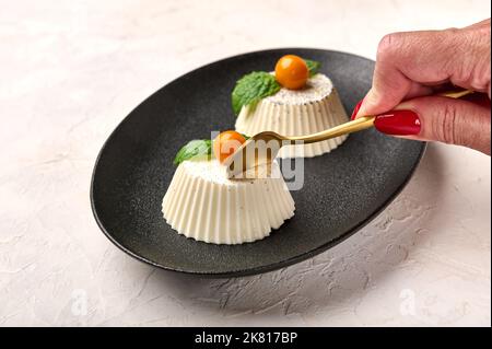 Die Hand der Frau hält Löffel über hausgemachter Panna Cotta mit natürlicher Vanilleschote, Physalis oder Winterkirsche und Minze auf schwarzem Teller, selektiver Fokus Stockfoto