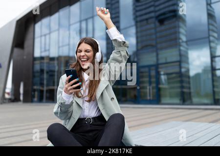 Junge stilvolle Mädchen hört Musik und tanzt mit den Armen, während sie vor dem Hintergrund der Fassade eines modernen Gebäudes sitzt Stockfoto