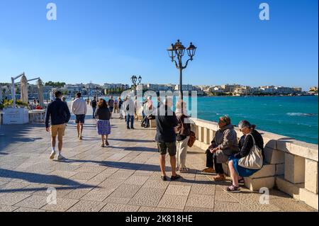 Touristen, die bei strahlendem Nachmittagssonne entlang der Uferpromenade von Otranto auf der Lungomare degli Eroi (die Heldenpromenade) spazieren. Apulien (Apulien), Italien. Stockfoto