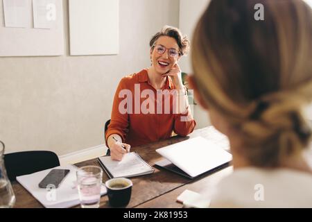 Weibliche einstellende Managerin lächelt fröhlich, während sie einen Kandidaten in ihrem Büro interviewt. Fröhliche Geschäftsfrau, die ein Treffen mit einem Job in der engeren Auswahl hat Stockfoto