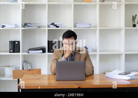 Asiatischer Geschäftsmann fühlt sich gestresst und frustriert, während er mit einem Laptop arbeitet. Männlicher Unternehmer immer in Schwierigkeiten mit seinem Job. Freiberuflich Stockfoto