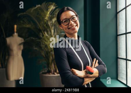 Porträt einer selbstbewussten Modedesignerin, die mit gekreuzten Armen steht. Junge Schneiderin lächelt in ihrem Modestudio vor der Kamera. Stockfoto