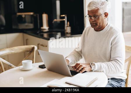 Ein reifer Geschäftsmann tippt auf einem Laptop, während er in seinem Heimbüro sitzt. Leitender Geschäftsmann sendet E-Mails und erstellt Geschäftspläne, während er von aus arbeitet Stockfoto