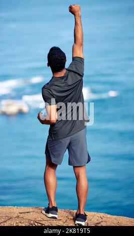 Fit und frei ist das Hauptziel. Rückansicht eines sportlichen jungen Mannes, der beim Training entlang der Küste mit ausgestreckten Armen steht. Stockfoto