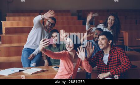 Fröhliche Schüler haben Spaß, lustige Videos im Klassenzimmer aufzunehmen, winkende Hände zu winken und lächelnd an Schreibtischen im Hörsaal zu sitzen. Selfie, Smartphone und Bildungskonzept. Stockfoto